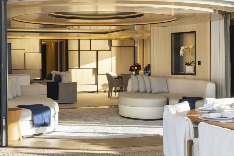 Luxurious cream-colored sofas and a dining table with white-covered chairs fill the foreground, while the background reveals a modern living space with geometric wall paneling and a circular ceiling design.