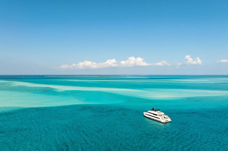 A white yacht floats on the clear turquoise waters of a shallow sea, with a bright blue sky and distant clouds in the background.