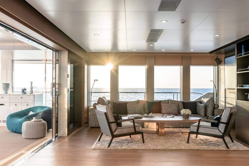 Sunlight streams into a luxurious yacht lounge with comfortable seating in the foreground and a serene ocean view through large windows in the background.