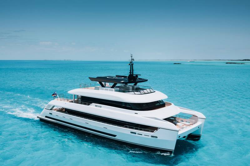 A sleek white catamaran yacht glides through crystal-clear turquoise waters under a vast, cloudless blue sky.