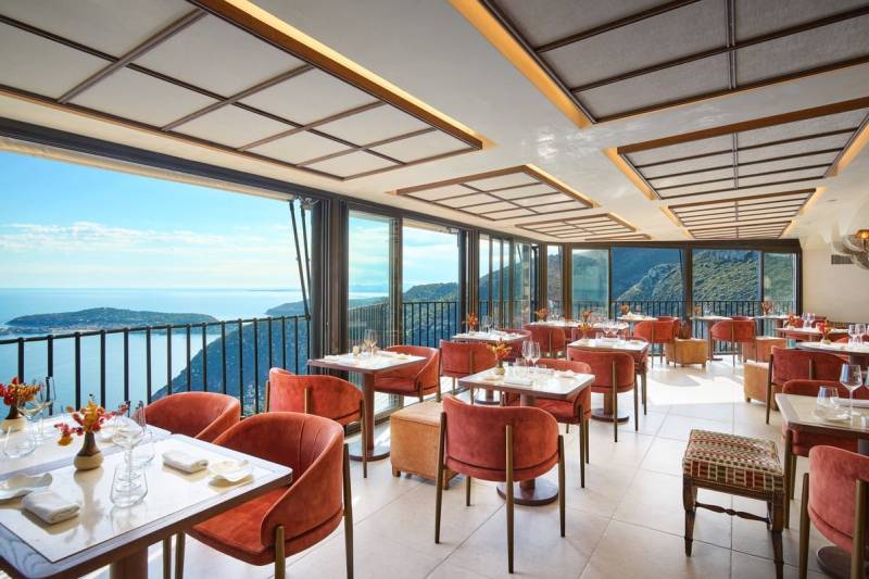 Elegant dining tables with orange chairs fill the foreground of a restaurant with a breathtaking ocean and mountain view in the background.