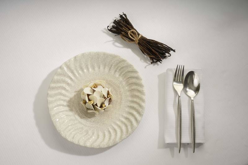 A white textured plate with a small dessert sits on a white surface, with a bundle of vanilla beans and silverware to the right.