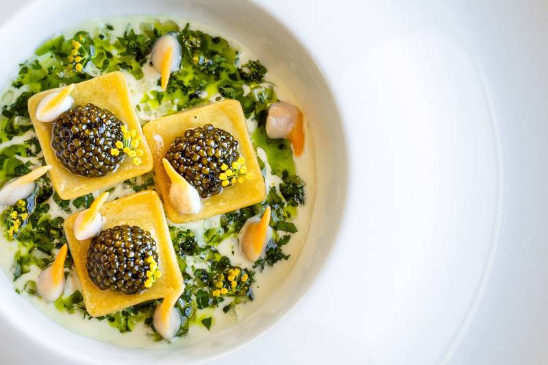 Three square ravioli topped with caviar are served in a creamy green sauce with scattered yellow garnishes, set against a blurred background of a white bowl and a warm, neutral surface.