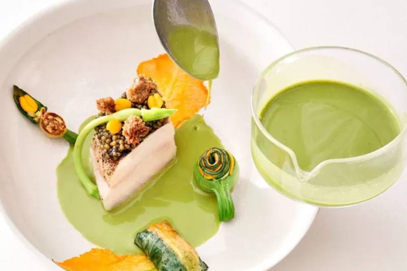 A spoon pours green sauce onto a meticulously plated dish of fish and vegetables, with a small glass of the same sauce beside it.