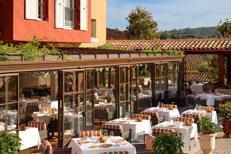 An outdoor dining area with tables set for meals is in the foreground, while a vibrant orange building and tiled roofs form the background.
