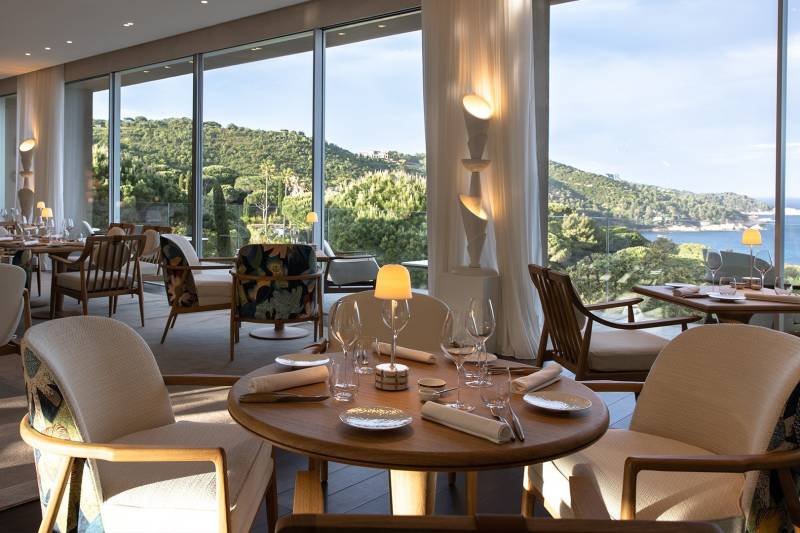 A warmly lit dining table in the foreground is set for a meal, with a panoramic view of a sun-drenched coastline and ocean visible through large windows in the background.
