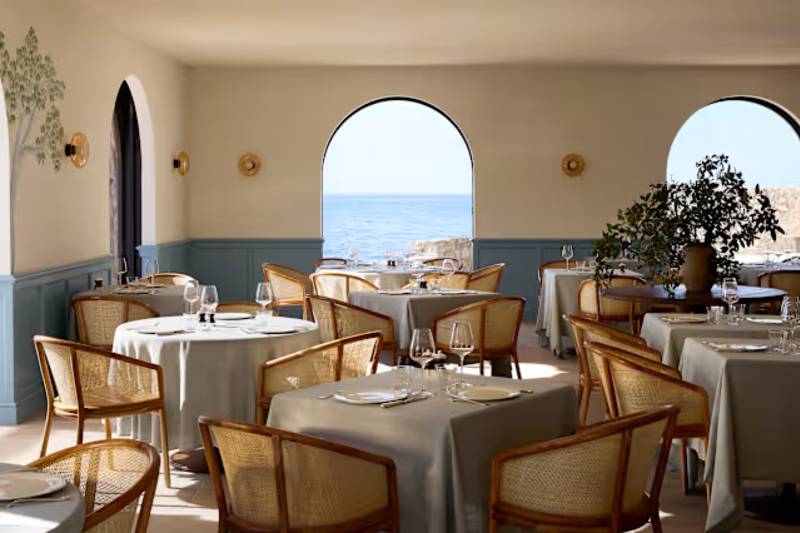 Rattan-backed chairs surround tables set with white tablecloths in a bright restaurant, with arched windows in the background revealing a view of the blue ocean.