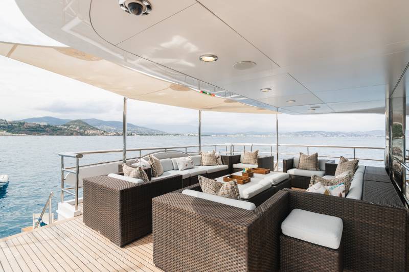 A spacious, covered outdoor lounge area with wicker furniture and plush cushions sits on a yacht's deck, overlooking a calm sea and a distant, hilly coastline.