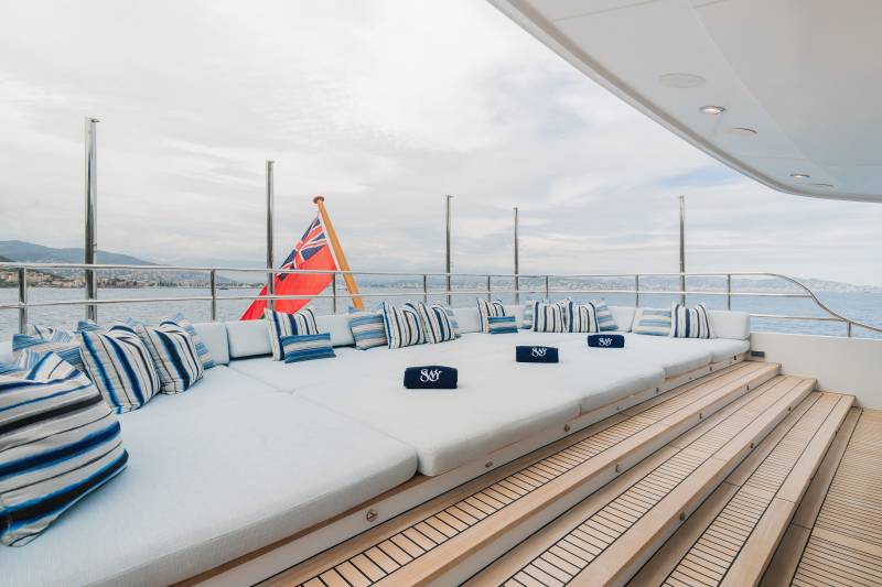 A spacious, white cushioned deck with blue and white striped pillows and a British flag in the foreground overlooks a calm sea and distant coastline in the background.