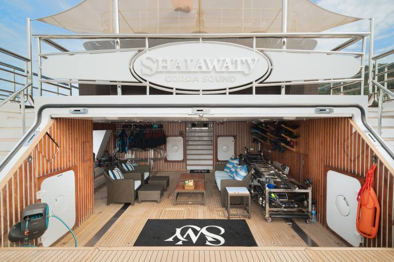 A spacious, open-air deck of a yacht, featuring comfortable seating and water sports equipment in the foreground, with the yacht's name "SHAFATY" prominently displayed on the upper deck in the background.