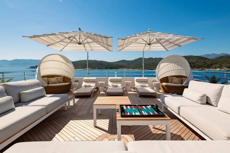 White sofas and lounge chairs with umbrellas are arranged on a wooden deck in the foreground, while a calm blue sea and green hills are visible in the background.