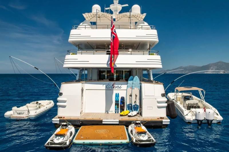 A large white yacht with watercraft and water toys in the foreground floats on a calm blue sea with a distant mountainous coastline in the background.