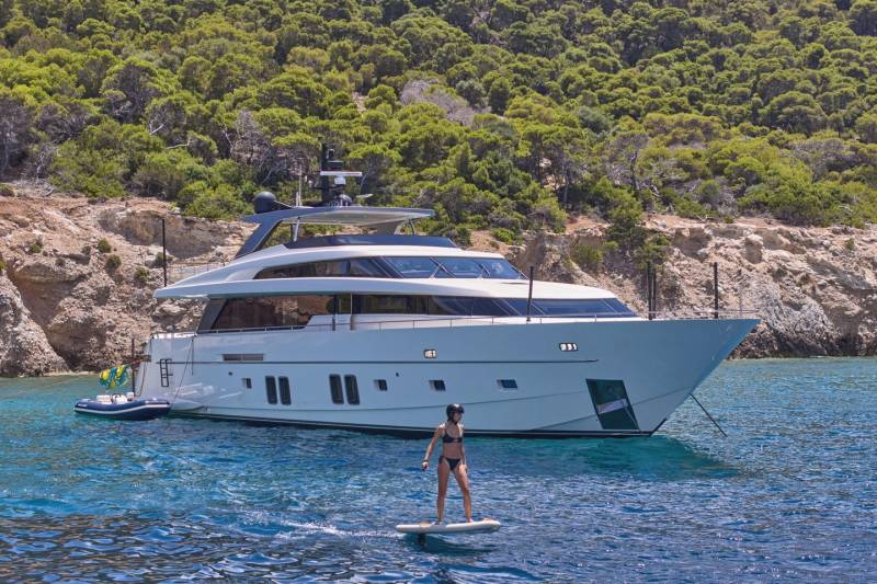 A woman on a paddleboard glides on clear blue water in front of a large white yacht anchored near a lush, green, rocky coastline.