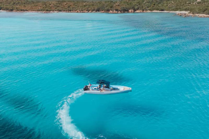 A white speedboat leaves a swirling white wake in the crystal-clear turquoise water of a bay, with a tree-covered coastline and distant mountains under a bright sky in the background.