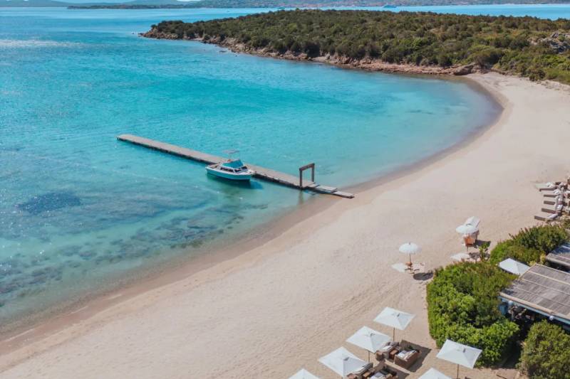 White umbrellas and lounge chairs dot a sandy beach in the foreground, leading to a calm turquoise bay with a wooden pier and a boat, all set against a backdrop of lush green hills under a clear blue sky.