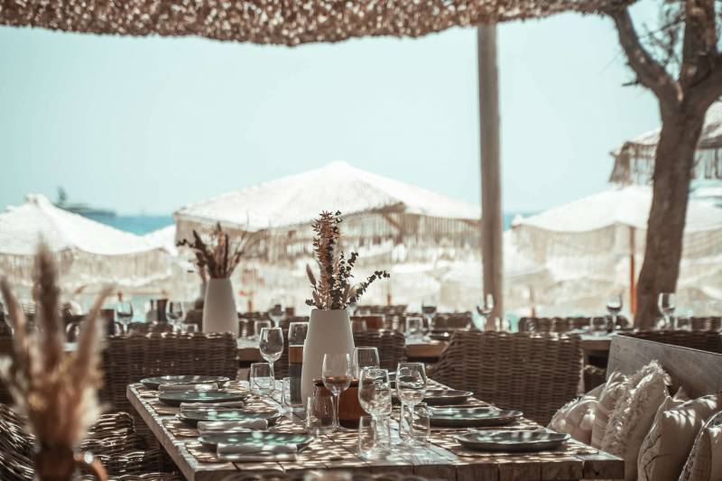 A beautifully set outdoor dining table with woven chairs and floral arrangements in the foreground is set against a backdrop of white beach umbrellas and the ocean under a thatched roof.