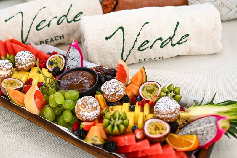 A vibrant fruit platter with pastries sits on a white surface in front of plush pillows and towels embroidered with "Verde Beach."