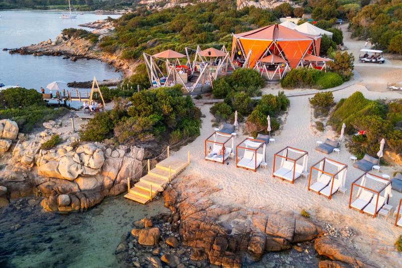 In the foreground, clear turquoise water laps against a rocky shore dotted with white cabanas, while the background features a vibrant orange building nestled amongst lush green foliage and rolling hills under a cloudy sky.