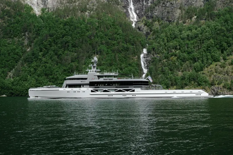 A sleek, modern yacht cruises on calm water in the foreground, with a lush, green mountainside featuring a cascading waterfall in the background.