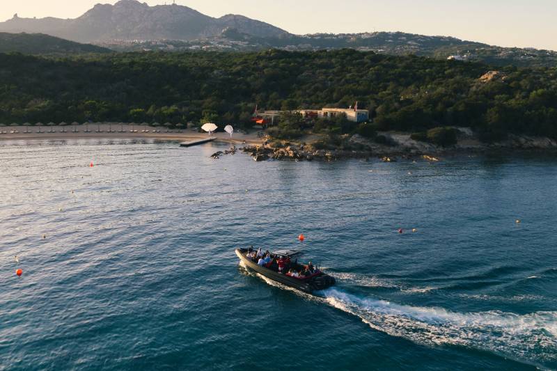 A boat with people on board cruises across the blue water in the foreground, with a tree-covered coastline and hills in the background.