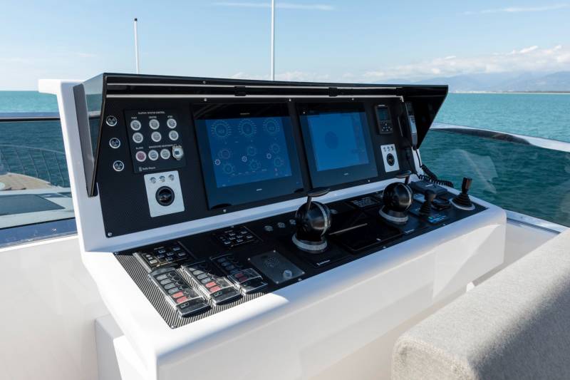 A modern yacht's helm station with multiple screens and controls is set against a backdrop of calm blue water and distant mountains under a clear sky.