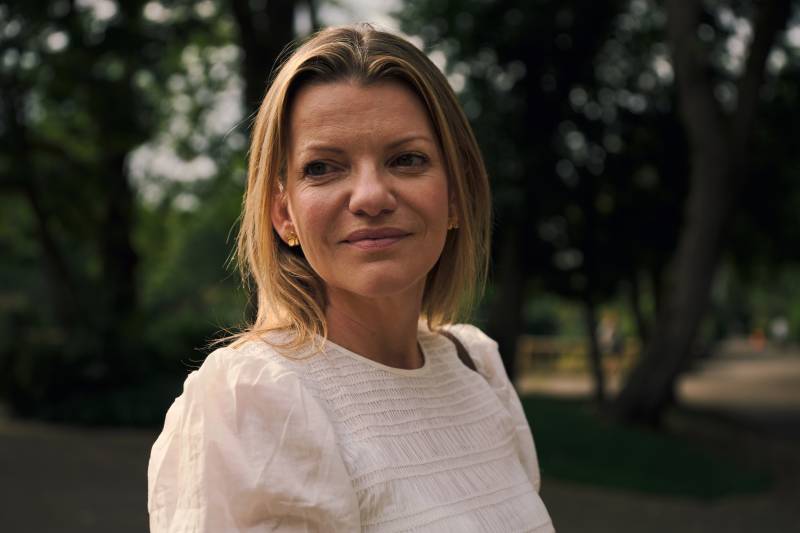 A blonde woman in a white top smiles softly in the foreground, with a blurred background of trees and foliage.