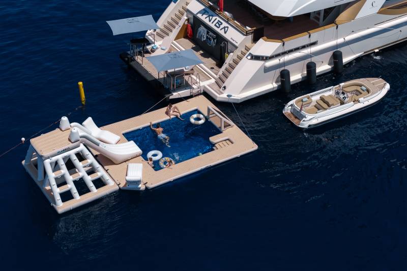 A floating pool with people swimming and a slide is anchored to a large yacht, with a smaller boat docked nearby in the deep blue water.