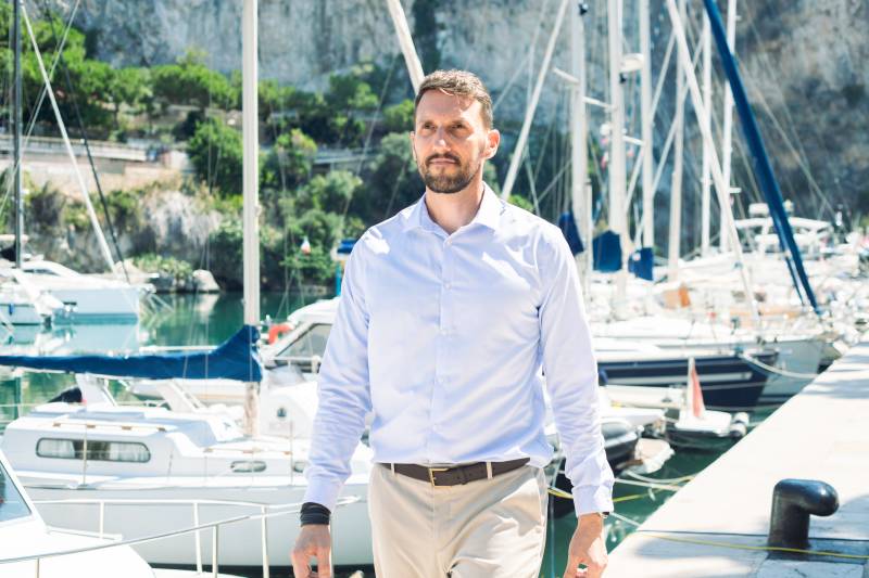 A man in a light blue shirt and tan pants walks along a dock with sailboats and a rocky cliff in the background.