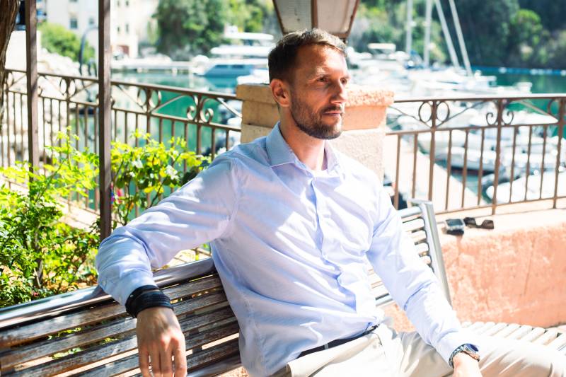 A man in a light blue shirt and beige pants sits on a bench in the foreground, with a harbor full of boats and lush greenery in the background.