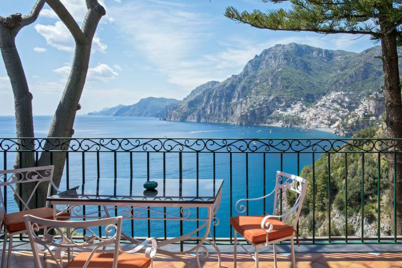 A table and chairs on a balcony overlook a sparkling blue sea and a picturesque coastal town nestled among green mountains.