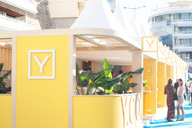 Bright yellow booths with a "Y" logo line a blue carpeted walkway, with people milling about and buildings in the background.