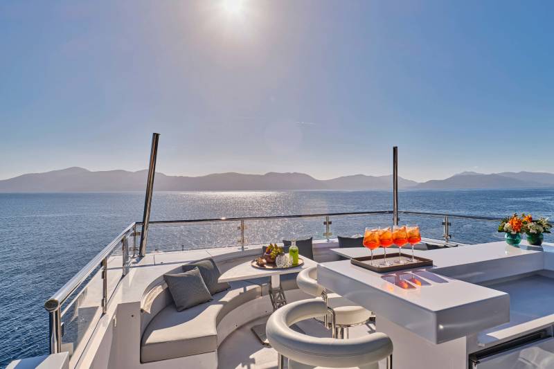 A luxurious yacht deck with a curved sofa and bar in the foreground overlooks a serene ocean and distant mountains under a bright sun.