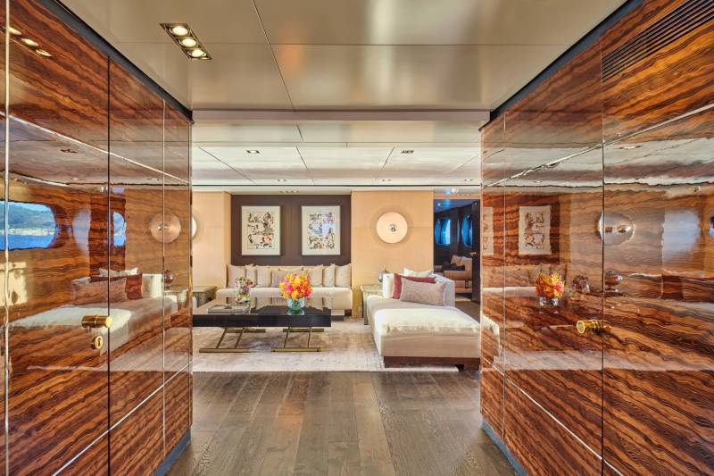 Polished wood paneling lines the foreground hallway, opening to a well-lit, modern living area with plush seating and artwork in the background.