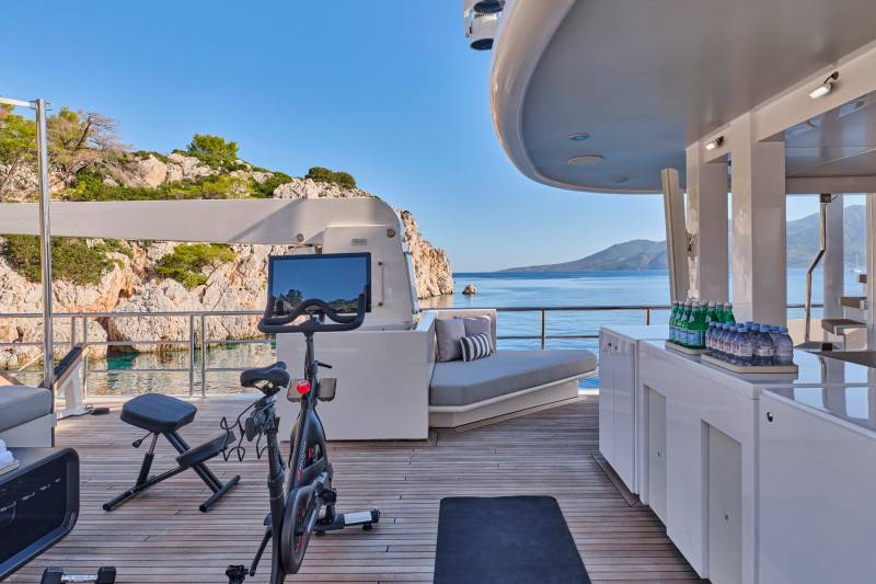 A Peloton bike and weights are set up on a yacht's deck with a scenic coastal view in the background.