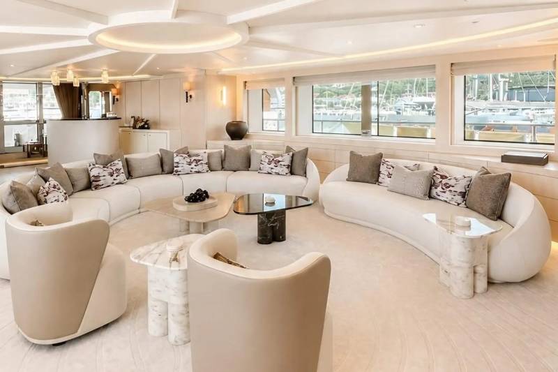 A luxurious, curved cream sofa and modern coffee tables fill the foreground of a bright yacht interior, while large windows in the background reveal a serene view of boats docked in a harbor.