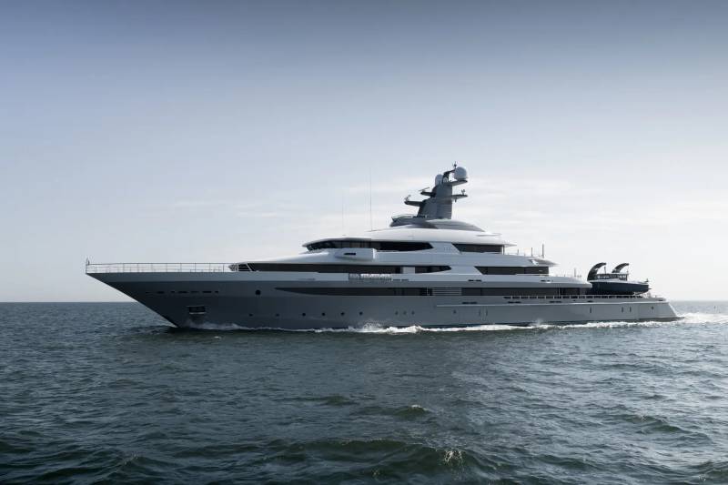 A sleek, gray superyacht cruises on calm, dark blue water under a pale, hazy sky.