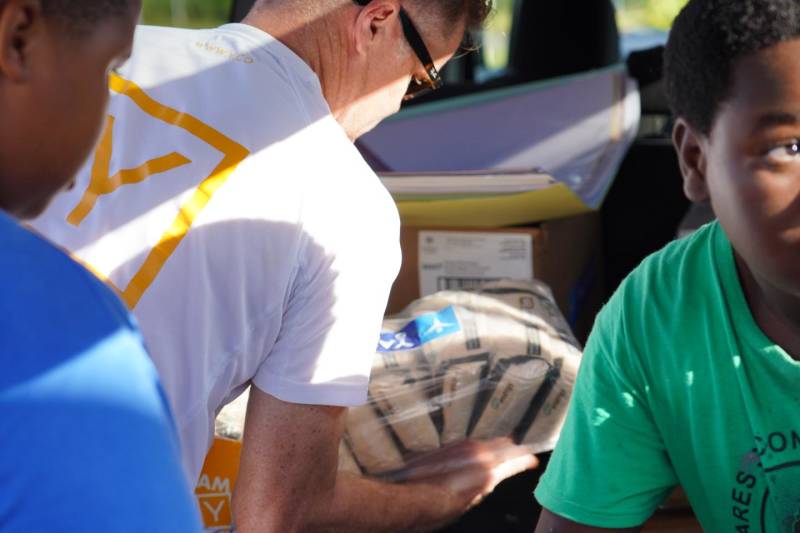 In the foreground, two young boys in blue and green shirts look on as a man in a white shirt holds a package, while the background shows the interior of a vehicle filled with boxes.