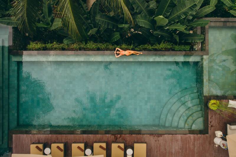A woman sunbathes by a turquoise pool surrounded by lush green foliage and lounge chairs.