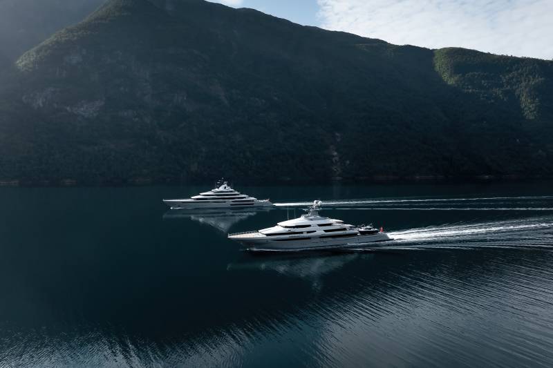 Two sleek yachts glide across a calm, dark blue body of water, leaving white wakes, with a majestic, tree-covered mountain range rising in the background under a hazy sky.