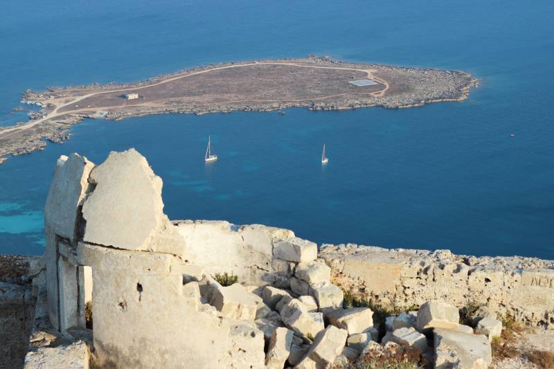 Crumbling stone ruins in the foreground overlook a tranquil blue sea with two sailboats and a distant, arid island.