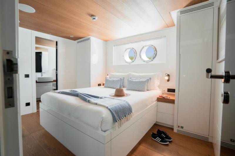 A neatly made bed with blue and white striped pillows and a straw hat sits in the foreground of a bright, modern cabin with porthole windows in the background.