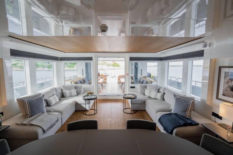 Plush sectional sofas and small tables fill the foreground of a luxurious yacht's lounge, while large windows reveal a serene waterfront view in the background.