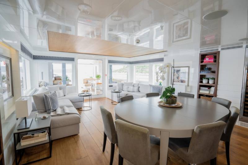 A round dining table with upholstered chairs sits in the foreground, while a spacious living area with a sectional sofa and large windows fills the background.