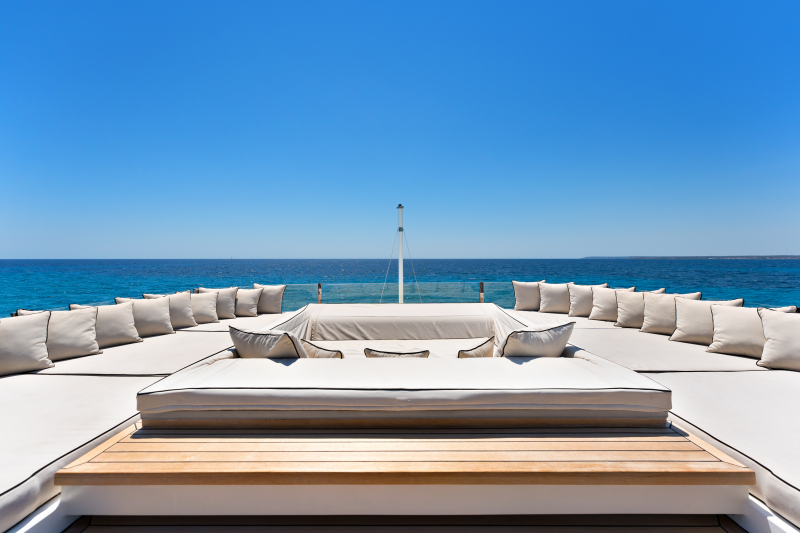 Plush white cushions arranged on a wooden deck face a vast expanse of blue ocean under a clear sky.