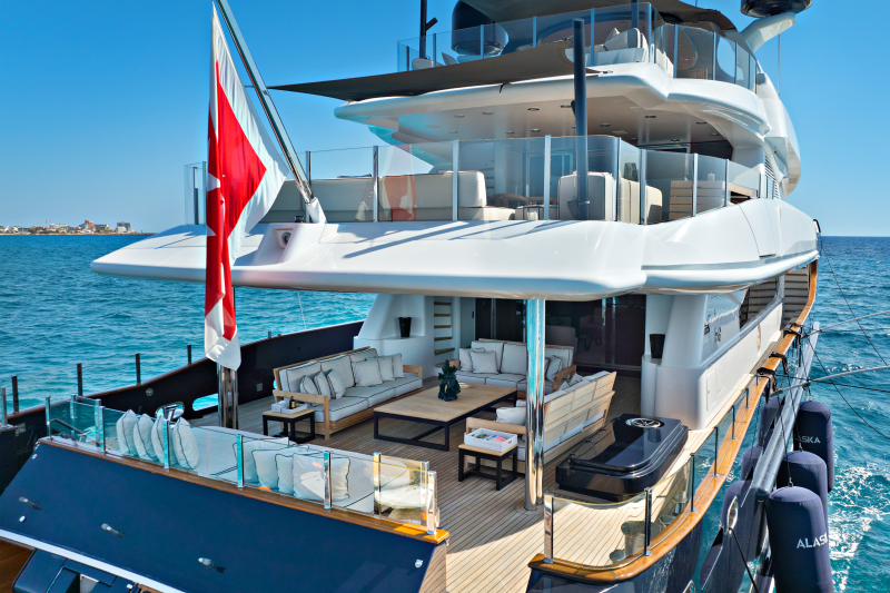 A luxurious yacht with a red and white flag flies in the foreground, while a calm blue sea and distant coastline form the background.