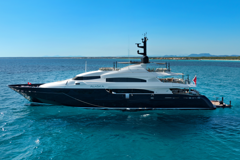 A large, dark blue and white yacht named "Alaska" floats on calm, turquoise water with a distant, hazy coastline under a clear blue sky.