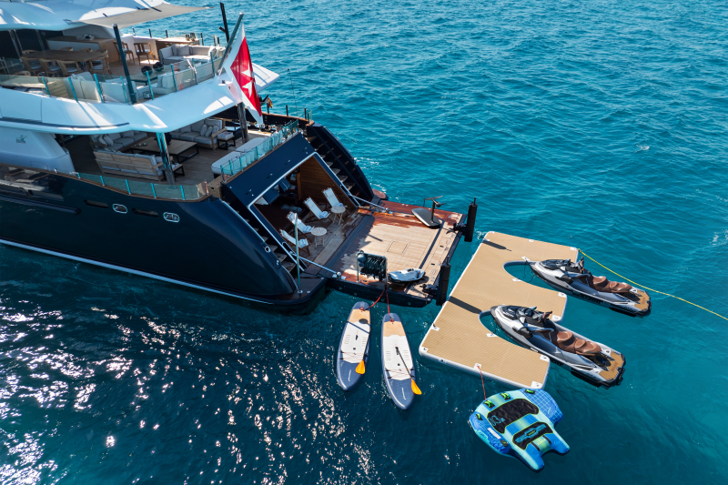 A luxurious yacht with a swim platform, jet skis, paddleboards, and an inflatable water toy floats on the clear blue ocean.