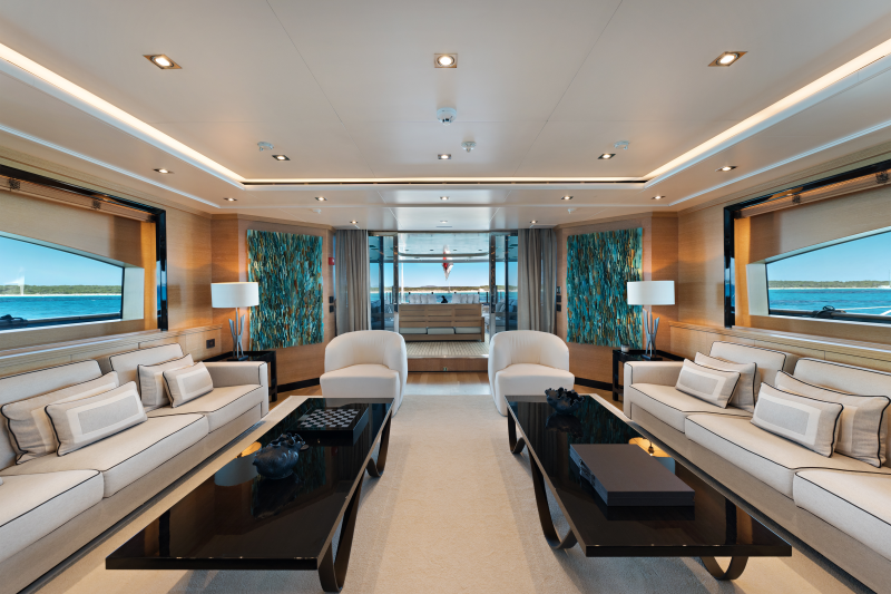 Luxurious cream sofas and dark coffee tables fill the foreground of a yacht's lounge, while the background reveals an open doorway with a view of the ocean.