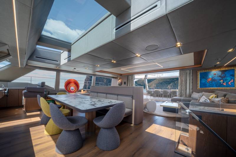 A modern yacht's dining area with colorful chairs and a patterned table is in the foreground, while a spacious lounge with a large artwork and ocean views is in the background.