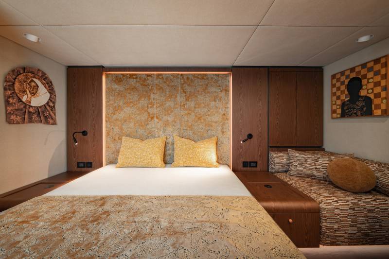 A cozy bedroom features a neatly made bed with yellow pillows in the foreground, while the background reveals a textured wall, wooden cabinets, and two framed artworks.